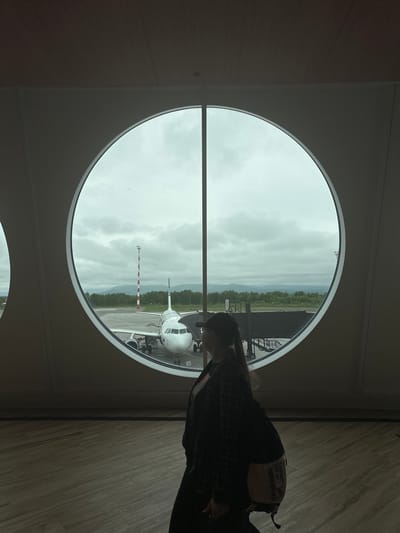 The image captures a view from inside an airport terminal in Yuzhnyye Koryaki, Russia. A young woman, silhouetted from the back, stands with her side to the camera. She has her hair pulled back in a ponytail and is wearing a dark plaid jacket over a light-colored top, with a backpack slung over her shoulder. She appears to be looking out of a large, circular window.
Through the window, an airplane is parked on the tarmac. The plane is white with a blue tail. A jet bridge is connected to the plane, and a tall red and white striped tower stands nearby. The background reveals a landscape of trees and mountains under a cloudy sky. The interior of the terminal is modern, with a wooden floor and white walls. The lighting is somewhat dim, with the light from the window being the primary source of illumination.