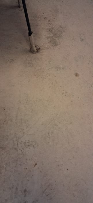 The scene depicts a light-colored concrete or tiled floor in Drugan, Bulgaria. A dark, slender cylindrical pole extends from the upper left downward towards the center-left. The base of this pole is wrapped in what appears to be light beige or off-white adhesive tape, forming an irregular, roughly conical foot approximately 5-7 cm in diameter at its widest point. The floor surface exhibits multiple scuff marks, dark smudges, and areas of discoloration consistent with dirt and wear. In the middle-right section, faint quadruped paw prints are visible. A small, distinct dark brown spot is located in the upper right quadrant of the image. No individuals or active interactions are present within the frame.