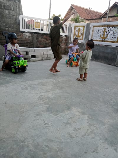 The image shows four children playing on a concrete surface in Bungur, Indonesia. A child sits on a green toy tractor, wearing a helmet labeled "Police." Another child stands barefoot with an arm raised. A third child rides a smaller, orange and green toy vehicle. The fourth child, wearing sandals and a striped outfit, stands observing. A low brick wall with a white decorative fence borders the area, with houses visible in the background. The lighting suggests it is daytime, likely late afternoon or early evening.