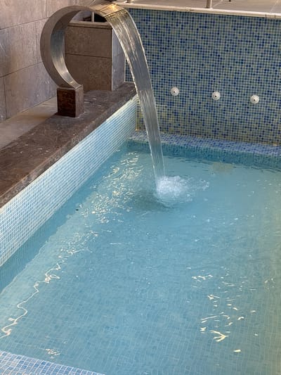 A small, square pool is filled with clear, blue water. A thin stream of water pours from a chrome fixture, creating ripples and bubbles on the surface of the pool. The pool is surrounded by a tiled wall. To the left of the pool is a decorative metal sculpture and a stone ledge. The scene is an outdoor setting, likely a spa or garden area, in Ayn al Basha, Jordan.