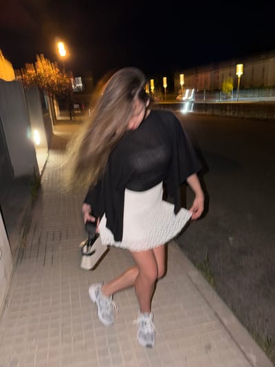 A blurry, rotated photograph shows a person sitting on a sidewalk at night. The person has long blonde hair and is wearing a black top and a white dress. They are wearing white sneakers. The sidewalk is made of gray bricks. To the left of the sidewalk is a dark area, and further back, blurred streetlights and buildings are visible. The location is possibly Platja D'alcúdia, Spain, given the information provided.