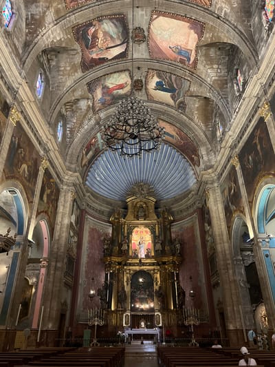 The image shows the interior of a church, likely in Pollença, Spain, characterized by its ornate architecture and religious art. The view is from the nave, looking towards the altar. The ceiling features a vaulted structure adorned with painted murals depicting religious scenes. A large, elaborate chandelier hangs from the center of the ceiling. The walls are decorated with more paintings and gilded architectural elements. The main altar area is highly decorated with statues and a central religious icon, framed by a golden retablo. Rows of wooden pews are visible in the foreground, with a few individuals seated and others walking in the background near the altar. The lighting appears to be a combination of natural light filtering through stained glass windows and artificial lighting from fixtures and candles. The overall impression is one of grandeur and historical significance.