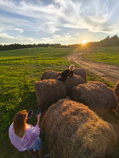 The image depicts an outdoor scene at sunset in a rural landscape, likely Стеклянный, Russia. In the mid-ground, a woman with blonde hair, dressed in a black long-sleeved top and dark pants, is posing on a stack of large, round hay bales. She reclines on one bale while another woman, positioned in the foreground with her back to the viewer, is actively photographing her. This photographer has long brown hair and is wearing a light pink, possibly satin, long-sleeved top and denim shorts. She holds two smartphones, both with dark screens, up towards the subject.

The setting is an expansive green field, illuminated by the warm, golden light of the setting sun. The sun is visible on the horizon, partially obscured by a line of dark green trees that stretch across the background. A dirt path or road winds through the field towards the distant tree line. The sky above is a blend of light blue and soft white, with wispy cirrus clouds adding texture. The hay bales are bathed in the golden hour light, highlighting their straw textures. The overall atmosphere is serene and vibrant, capturing a moment of photography amidst the beauty of the countryside during dusk. No visible text is present in the image.