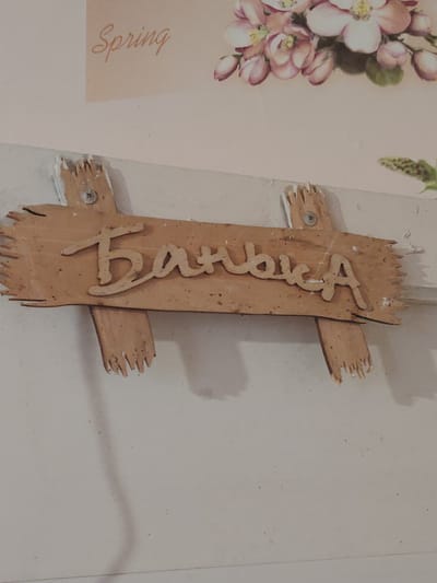 The image depicts a close-up, slightly angled view of a wooden sign that reads "Баня" in Cyrillic script, which translates to "Banya" or "Bathhouse" in English. The sign is made of light-colored wood and has a rustic, distressed appearance with frayed edges. It is attached to a white wall with visible screws and what appears to be some sort of white adhesive or plaster. Above the sign, a portion of a decorative print is visible, featuring the word "Spring" in cursive English and an illustration of pink blossoms. The overall environment suggests an interior setting, possibly a rural or traditional dwelling. There are no people or animals present in the frame. The lighting is soft, and the focus is on the sign itself. There is no information in the image to suggest a specific location in the city of Степное, Russia.