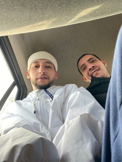 This image displays two adult males seated within a vehicle, captured from a low-angle perspective looking upward. The scene is situated in La Wantzenau, France.
The individual positioned on the left side of the frame has light skin, a short beard, and a mustache. He is wearing a light-colored, ribbed beanie and a white zippered jacket with a blue drawstring detail. His eyes are open, and he is looking directly at the camera with a neutral expression. His upper torso and head are prominently featured.
The second individual, on the right, is partially obscured by the first subject and a dark blue fabric visible on the extreme right. This male also has light skin and short, dark hair. He is smiling, revealing his teeth, and his gaze is directed towards the camera. He wears a dark-colored jacket, possibly black, with a visible textured collar or trim.
The background consists of the light grey, upholstered ceiling of a vehicle interior. A window frame is visible to the left of the individual on the left, indicating external daylight. No discernible details of the exterior are present. The overall lighting suggests daytime conditions.