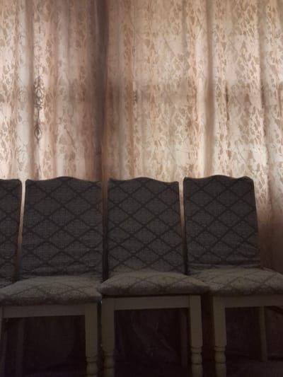 The image shows a row of four upholstered chairs against a patterned curtain. The chairs have light-colored wooden legs and the upholstery features a dark grey diamond pattern on the seat and a similar pattern on the back. The curtain behind the chairs is a light beige color with a subtle floral or leaf-like pattern. The lighting is dim, casting shadows on the floor in front of the chairs. The scene appears to be indoors, likely in a residential setting. There is no indication of the specific location within Қанабек, Kazakhstan.
