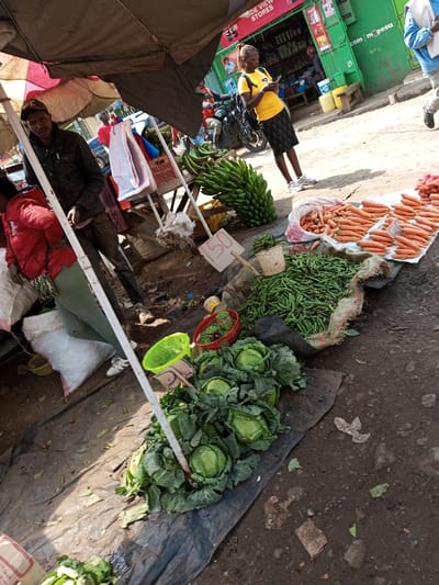 The image depicts an active outdoor market scene situated in Keekonyokie ward, Kenya. The foreground features a market stall arranged on uneven dirt ground under a dark tarp canopy. On a black tarp, several heads of fresh green cabbage are displayed. Adjacent to this, a large quantity of green beans or peas is piled on a woven burlap sack, alongside a small red woven basket and a bright green plastic basket containing smaller produce. To the right, multiple piles of orange carrots are arranged, some contained within a white sack, on a lighter-colored surface. A large bunch of green bananas rests on a wooden crate, with a white sign displaying "150" nearby.
Three individuals are prominent:
1. On the left, a person wearing a red jacket and green trousers is partially visible, standing next to large white sacks.
2. In the mid-ground, a man wearing a dark jacket, dark trousers, and a baseball cap stands looking forward.
3. To the right, a woman in a yellow short-sleeved top, a black patterned skirt, and white sneakers stands, holding and viewing a mobile phone. She is positioned in front of a green storefront labeled "DE VIEW STORES" with "m-pesa" branding. A motorcycle is parked behind her.
Another person, partially seen in blue and white attire, walks in the far background right. Additional vehicles, including a car, are visible in the background. The scene is brightly lit by natural sunlight, casting shadows from the canopy and market structures. The ground throughout the market area is dirt with scattered debris.