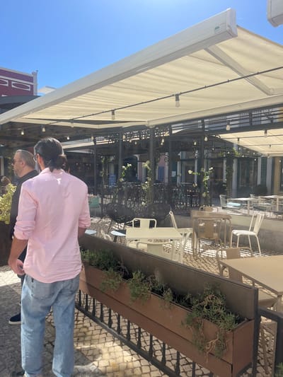 The image depicts an outdoor cafe terrace in Alcochete, Portugal, under a clear blue sky with bright sunlight. In the foreground, a male subject with dark hair tied back, wearing a light pink long-sleeved shirt and light blue jeans, stands facing away from the camera. To his left, another male subject with silver-rimmed glasses and a dark shirt is partially visible. Both are on a cobblestone surface.
A dark metal planter box, containing small green plants and mounted on a metal railing, delineates the cafe seating area. This area is shaded by a large, cream-colored fabric awning supported by a dark metal framework, which also holds multiple exposed bulb string lights. Numerous unoccupied tables and chairs, in white, beige, and black metal, are arranged on the cobblestone ground. Decorative black metal structures entwined with white flowering plants are present within the seating area.
In the background, buildings with traditional Portuguese architecture feature colored facades, including a reddish-purple upper section with white trim on the left, and other light-toned structures with blue window details.