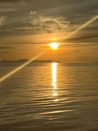 The image depicts a serene ocean sunset with a clear horizon. The sun is positioned in the upper center of the frame, casting a brilliant golden glow across the water and sky. A prominent sunbeam or lens flare streaks diagonally from the top right to the bottom left of the image. The water is calm with gentle ripples reflecting the sunset's colors. In the distance, a silhouetted landmass, likely islands or a coastline, is visible against the horizon. The sky is a blend of warm oranges, yellows, and muted grays, with wispy clouds scattered across it. There are no people or specific objects to note other than the natural elements of the seascape. The location is identified as Baan Pang Ka, Thailand, suggesting this is a scene from that coastal area.