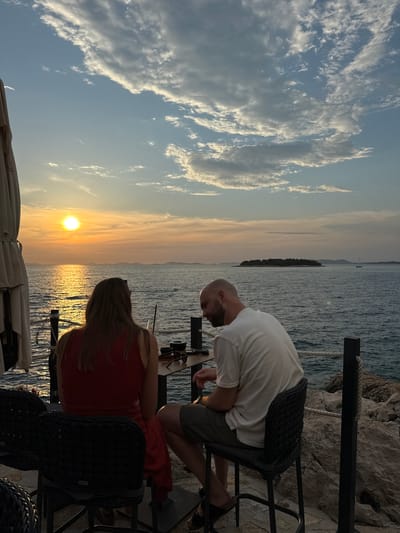 Two people, a man and a woman, are seated at a table by the sea in Primošten, Croatia, at sunset. The woman, facing away from the camera, wears a red dress. The man, also facing away from the camera, wears a white t-shirt and shorts. They are seated on chairs at a table with drinks. The sun is setting over the sea, casting a warm glow on the water. Islands and a sailboat are visible in the distance. The sky is filled with clouds. The scene appears to be at an outdoor restaurant or bar with a view of the Adriatic Sea.