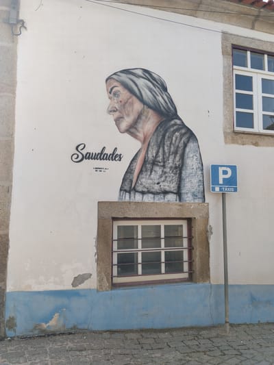 The image shows a building in São João da Pesqueira, Portugal, featuring a mural of an elderly woman with the word "Saudades" written above. The mural is located on the side of a building with two windows. A blue and white parking sign labeled "Táxis" stands near one of the windows. The building's exterior is marked by some weathering and discoloration. The ground is paved with cobblestones.