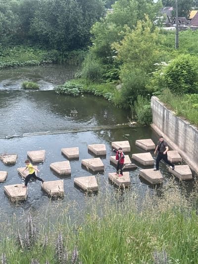 The image depicts a group of three people navigating a river in Markidonovka, Russia, by stepping on large, flat, square concrete blocks that protrude from the water. On the left, a young boy wearing a yellow t-shirt and black pants is mid-stride, jumping from one block to another. In the center, a child in a red jacket and black pants stands on a block, facing left and appearing to pose. To the right, an adult male, wearing a camouflage t-shirt and black pants, is actively walking across the blocks, looking to the left. The river is calm with some ripples around the blocks. The banks of the river are overgrown with lush green vegetation, including tall grasses and trees. A concrete wall runs along the right bank of the river, with more dense foliage behind it. In the background, there are glimpses of rural buildings with pitched roofs, typical of a village setting. The overall scene suggests a recreational activity on a summer day in a natural environment within the town.