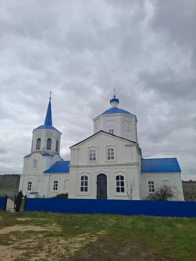 An exterior, daytime shot depicts a white Orthodox Christian church complex in Yablonovo, Russia, set against an overcast, cloudy sky. The main structure is characterized by its white facade, featuring multiple rectangular windows, some with arched tops, and dark, possibly wooden, doors on the lower level. Distinctive blue roofs and domes crown the buildings.

To the right, a classic blue onion dome topped with a cross is visible. To the left, a taller, conical blue spire, also adorned with a cross, rises from a separate tower-like section of the complex. The architecture is typical of Russian Orthodox churches.

A bright blue corrugated metal fence runs across the foreground, partially obscuring the lower sections of the church and separating it from a grassy and dirt area on the far right. A bare tree or bush is visible behind the fence, next to the building. In the distance, low hills can be seen under the heavy cloud cover. The sky is uniformly grey, indicating an overcast day without direct sunlight. There are no people visible, and no specific activity or event is taking place, presenting a static view of the architectural landmark. No visible text is present in the image.