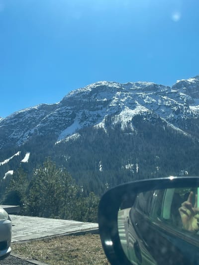 The image shows a landscape in the Sappada/Plodn/Sapade, Italy area, taken from inside a vehicle. A mountain range covered in snow and trees occupies the left and center of the frame, with a clear blue sky overhead. The right side of the image shows part of the vehicle's side mirror reflecting a hand and a portion of the interior. The foreground includes a roadside area with grass and a concrete barrier.