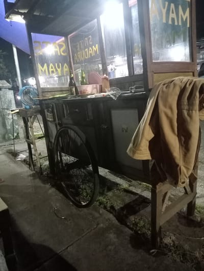 A food cart, apparently offering sate ayam (chicken satay) madura, is located on the side of a street in Blitaran, Indonesia. The cart has a bicycle wheel attached, suggesting mobility. A blue electric fan sits atop a makeshift table next to the cart. A tan jacket is draped over a wooden chair. The scene is dimly lit, suggesting it is nighttime. The cart is parked on a concrete sidewalk next to a patch of grass. Other buildings are visible in the background, slightly out of focus.
