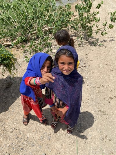 Three young girls are positioned outdoors on a gravelly surface, likely a dirt road. The two girls in the foreground are partially obscured by their traditional attire, with one wearing a bright blue headscarf and red patterned clothing, while the other is draped in a blue striped shawl over striped clothing. Both are wearing sandals. The girl to the left is pointing towards the viewer with her hand. A third girl stands partially hidden behind them, closer to a bush or small tree with green foliage. The setting appears to be rural, with sparse vegetation in the background.