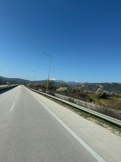The image displays a multi-lane asphalt highway under a clear blue sky, located within the Pamvotida Municipal Unit, Greece. The perspective is from the road, looking forward in the direction of travel.

The highway features a grey asphalt surface with a solid white line marking the right edge of the main lane and a dashed white line separating the two visible lanes. Minor surface cracking is evident on the asphalt. Along the right side of the highway, a metallic W-beam guardrail, supported by dark posts, runs parallel to the road. Beyond the guardrail, the terrain slopes downwards, revealing sparse vegetation, bare deciduous trees, and numerous trees adorned with white blossoms. Further to the right, a cultivated field of brown earth and a rocky hill covered in green vegetation are discernible. A series of tall, grey streetlights, each with a single luminaire, are spaced along the right edge of the road, extending into the distance.

In the mid-distance on the left lane, a white semi-truck with a trailer is visible, traveling in the same direction. A smaller, dark vehicle is positioned further ahead on the road. The background features a clear, uniformly blue sky above a distant mountain range, which includes two prominent snow-capped peaks rising above lower, greener hills. No individuals are visible in the scene.