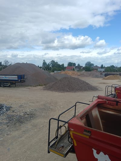 The image presents an industrial site in Яльгелево, Russia, likely a materials yard. The scene is dominated by several large piles of aggregate materials, including gravel, sand, and stones, of varying colors ranging from gray to tan. In the background, a blue and white truck with the inscription "CTPOU BETOH" (Building Concrete) is parked near the largest pile. In the foreground, a portion of a red industrial vehicle, specifically its rear cargo container and access ladder, is visible. Scattered piles of broken stone slabs are present on the ground to the left. The terrain consists of compacted dirt and gravel. The sky is overcast with a mix of clouds and patches of blue. Houses and trees are visible in the distant background, marking the edge of the industrial area.