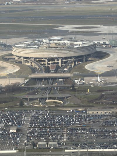 An aerial view showcases a circular airport terminal labeled "Terminal 1." Adjacent to it, runways stretch out, alongside an airplane parked at a gate. Roads and roundabouts weave around the terminal, connecting it to a large parking area filled with densely packed cars. Green spaces are interspersed throughout the area, providing contrast to the built environment.