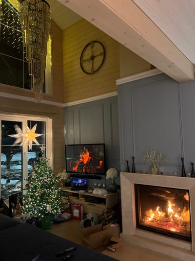 This image depicts a cozy and festive interior room, likely a living room, in Finskiy Briz, Russia, during the evening or night.
The room features a blend of architectural styles: the left side showcases a high, light-colored wooden-paneled ceiling with prominent wooden beams, suggesting an attic or sloped roof structure. A large, intricate chandelier with many sparkling elements hangs from this area. Further back on the wooden ceiling, a large, dark metal wheel-like decoration is mounted. Below the sloped ceiling, a window is decorated with a large, illuminated paper star.
The right side of the room presents a modern aesthetic with a flat blue-grey ceiling and matching walls adorned with decorative molding. A prominent fireplace with a light-colored stone surround is on the right wall, featuring a warm, crackling fire. The mantelpiece above the fireplace holds decorative items including tall black vases and a white, coral-like sculpture. To the left of the fireplace, a flat-screen television is mounted, displaying a vibrant image of a person with sparkling lights, possibly from a music video or festive program.
Below the television and extending into the mid-ground, a brightly lit Christmas tree stands, decorated with numerous lights and ornaments. Several wrapped presents are visible at its base. Adjacent to the TV and Christmas tree, an entertainment unit or shelving unit holds various objects, including some open cardboard boxes and additional wrapped gifts. A dark-colored sofa is partially visible in the lower right foreground.
The scene conveys a warm, inviting, and festive atmosphere, characteristic of holiday celebrations. The internal lights, the glow from the fireplace, and the decorated Christmas tree indicate it is nighttime. No visible text is clearly discernible in the image.