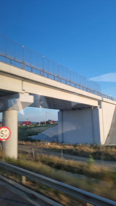 The image captures a view from a moving vehicle, likely on a highway, with a large overpass dominating the foreground. The overpass is constructed of concrete and has a metal railing along its edge, possibly for safety or to support a fence. Below the overpass, the landscape is rural, with dry, grassy fields, a dirt road, and several buildings with red roofs. Power lines and a tall pylon are visible in the distance. The sky is clear and blue. A circular traffic sign with the number "5" inside a red circle is attached to a concrete pillar on the left side of the image. The overall impression is one of infrastructure development in a semi-urban or peri-urban area of Schineni, Romania.