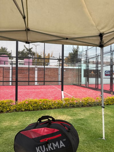 The image shows a view from under a light-colored tent structure, looking out at a padel tennis court. The court is red and enclosed by a black wire mesh fence and glass walls. Green shrubbery runs along the side of the court closest to the viewer. In the foreground, on the grass, is a black and red KUKMA brand sports bag. Reflections in the glass walls reveal buildings and trees outside the court area. San Francisco Totimehuacán, Mexico, is indicated by the metadata.