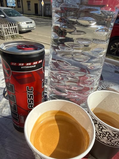 This image depicts an outdoor scene in Sarantsi, Bulgaria, under direct sunlight. In the foreground, resting on a light-colored surface, are three beverages: a red and black "BIG SHOCK CLASSIC" energy drink can (250ml) with a red pull tab, displaying "VITAMINS B2 B3 B5 B6 B12"; a tall, clear plastic bottle of water with a distinct rippled texture; and two white paper cups, each containing a liquid resembling espresso with visible crema. The water bottle exhibits reflections of the surrounding environment, including a red vehicle on the far right.

Beyond the immediate foreground, a low white metal fence separates the viewing point from a paved street. A silver Opel Astra sedan is parked on the left side of this street, with its rear section and a Bulgarian license plate partially visible. Further in the background, a light-colored building with dark windows is present. A yellow diamond-shaped priority road sign is mounted on a pole near the center-right of the background, and a blue and white directional sign is visible on the far left. No individuals are present in the frame.