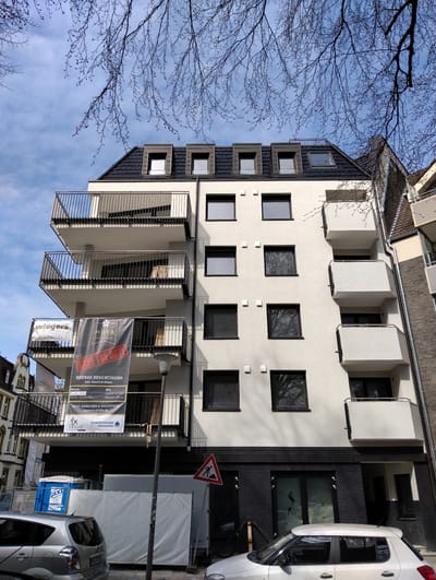 The image depicts a modern, multi-story residential building in Münster, Germany, as indicated by the German signage and the text "Hoyastraße 17." The building is white with black window frames and balconies. There are banners on one of the balconies advertising a "Neubau Besichtigung" (new building viewing) event scheduled for March 15, 2026. The banners feature the names "wiegers" and "HOYA." At the building's base, there's dark brick cladding, and signage with the text “isover” is visible behind what appear to be glass entryways. Construction or renovation activity is underway, evidenced by a portable toilet labeled "DIXI," construction barriers, and a warning sign indicating road work. Two parked silver/white cars are in front of the building. Branches from bare trees are visible in the upper portion of the image against a partly cloudy sky. The building is adjacent to another building, and a street lamp is also visible.