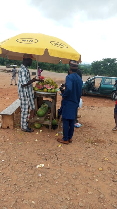 In Dauda, Nigeria, two men stand by a roadside fruit stand under a large yellow umbrella bearing the "MTN" logo. One man, wearing a checkered shirt and pants, is in the process of selecting a slice of watermelon from a bowl of assorted fruits. The other man, dressed in a blue tunic and pants, faces away from the viewer, observing the fruit selection. A wooden stand displays several watermelons on a lower shelf and a variety of fruits, including oranges and watermelons, on an upper shelf. In the background, a green vehicle with an open door is parked nearby. The ground is covered in dirt and scattered fruit peels. The scene appears to be a typical roadside vendor in a rural or semi-urban setting.