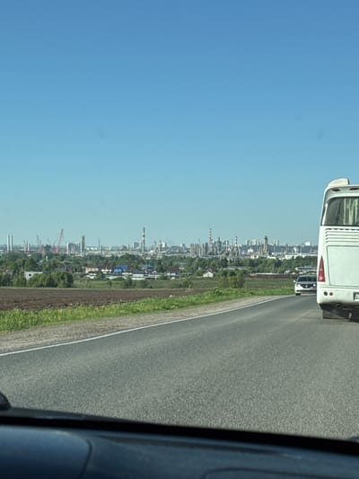 The image presents a view from inside a vehicle, potentially a car or bus, moving along a road. To the left, the sky is predominantly blue. On the right, the road stretches out, with a white dividing line visible. Beyond the road, there is a stretch of green vegetation. In the distance, a cityscape is visible, including buildings, trees, and industrial elements like cranes and smokestacks. The location is identified as Osentsy, Russia. A white bus is seen near the top of the frame. A portion of the interior of the vehicle from which the photo is taken can be seen on the right side.