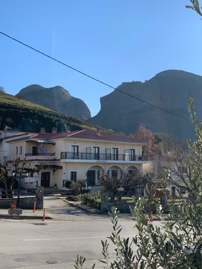 This image captures a vibrant daytime scene in Kastraki, Greece, showcasing a hotel building against a dramatic natural backdrop.
In the midground, a two-story hotel with a light yellow facade and a reddish-brown tiled roof is prominent on the left. It features several balconies adorned with dark, ornate metal railings. An arched section on the building's front clearly displays the name "HOTEL KOKOS" in dark letters. A Greek flag is proudly flown from a lower-level balcony. Surrounding the hotel are various trees and green foliage, some of which appear bare, suggesting a cooler season.
Towering in the background are the distinctive, imposing grey rock formations characteristic of the Meteora region. The sky above is clear and a brilliant blue, indicating bright, sunny weather. A dark utility line cuts diagonally across the upper left portion of the sky.
To the right, a paved road or street extends into the foreground. A couple of orange traffic cones are visible along its edge. A white sign near the hotel on the roadside displays text in Greek, which reads "PIZZA", "ΣΟΥΒΛΑΚΙ" (SOUVLAKI), and "ΚΡΕΠΕΣ" (CREPES). Olive tree branches, laden with green leaves, partially frame the scene from the upper and lower right corners. The overall impression is one of a serene, sunny day in a picturesque setting.