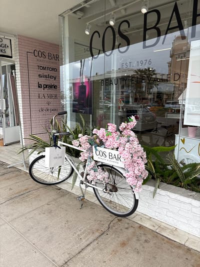 This image, rotated 90 degrees clockwise, captures an outdoor scene in Newport Beach, United States of America, centered on a beauty store storefront. A prominent feature is a white bicycle, heavily decorated with numerous clusters of pink artificial flowers in its front basket area, acting as a decorative display. A small white sign attached to the bicycle reads "COS BAR BEAUTY ELEVATED," and a white shopping bag with the "COS BAR" logo hangs from its handlebars.
The bicycle is positioned on a concrete sidewalk next to a building with large glass windows. The windows reflect the outdoor environment, including parts of a palm tree, other buildings, and vehicles. On the storefront's glass, reflections show "COS BAR," "EST. 1976," and "BEAUTY ELEVATED" written on the interior or another surface. Below the main glass windows, a section of light pink brick wall serves as additional signage, prominently featuring "COS BAR BEAUTY ELEVATED" and a list of luxury beauty brands: "TOM FORD," "sisley PARIS," "la prairie SWITZERLAND," "LA MER," and "clé de peau BEAUTE." Green foliage is visible near the base of the storefront and around the bicycle. The concrete sidewalk shows some wear and cracks. The bright lighting suggests it is daytime. No people are visible.
