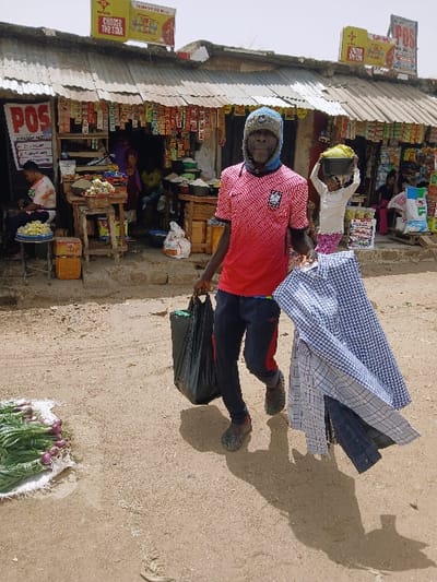 This image depicts a bustling outdoor market scene in Kadoma, Zimbabwe, under a bright sky. In the foreground, a dark-skinned adult male is walking towards the viewer. He wears a blue and grey knitted beanie, a red and white patterned short-sleeved shirt with an emblem on the left chest, and dark-colored track pants with red stripes. On his feet are dark, possibly muddy, closed-toe shoes. He carries a black plastic bag in his left hand and several folded, plaid, long-sleeved shirts in various shades of blue and white over his right forearm and hand. His shadow is cast prominently on the dry, light brown dirt ground. To his left, on the ground, a pile of green onions and purple onions rests on a white sack.

In the mid-ground and background, a row of small, informal market stalls or shops with corrugated metal roofs is visible. These structures are made of light-colored, possibly stucco or rough-hewn, walls. The storefronts display numerous packaged goods, including snacks and sachets, hanging from strings or stacked on shelves. Several "POS" (Point of Sale) signs are visible, one prominently on the left shop and another on the right. A yellow billboard with the text "MAGARA" and "CHOOSE THE STAR" is mounted above the central shop.

Inside and around the shops, multiple individuals are present. To the left, a person wearing a light-colored tie-dye shirt is seated, with a tray of yellow snacks on a blue stool nearby. Further right, a child with light clothing is visible, carrying a large bowl of green produce on their head. Other figures are indistinctly visible within the shaded interiors of the shops. The overall environment suggests a vibrant, open-air commercial area.