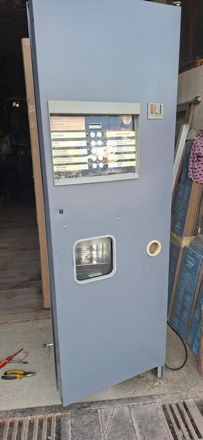 The image shows a large, grey, commercial vending machine standing upright on a concrete surface, likely outdoors or in a workshop. The machine has a control panel with buttons and a digital display, as well as various beverage options listed in Cyrillic and English. There is a dispenser opening on the front of the machine. Tools, including a screwdriver and pliers, are scattered on the ground near the base of the machine. In the background, stacks of blue boxes or packaging are visible, suggesting a storage or industrial setting. The scene is lit by natural light, and the overall impression is one of an outdoor or semi-outdoor workspace. The presence of Bulgarian text on the machine's interface suggests the location is in Bulgaria. The city of Dobrinishte, Bulgaria is known for its natural beauty and tourist attractions, and this vending machine could be a component of a public service or a business operating within the city.