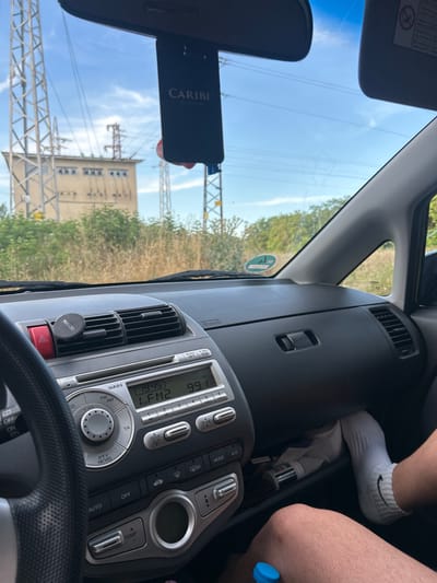 The image is a first-person perspective from inside a car, looking through the windshield. Outside the car, a large electrical substation is visible, with tall metal towers and power lines against a bright blue sky with scattered clouds. The interior of the car shows the dashboard, steering wheel, and car stereo. A black air freshener hangs from the rearview mirror. A person's legs, wearing white socks with a black Nike logo, are visible in the lower right corner of the frame, with a blue-capped bottle near the knee. The scene appears to be in an outdoor, possibly rural or industrial, setting.
