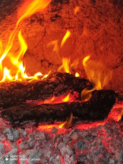 A vibrant scene shows logs ablaze in a fireplace. Bright yellow flames dance upwards, illuminating a rough, orange-red wall behind the logs. Beneath the logs, glowing red embers and gray ashes fill the space, indicating intense heat. The photograph captures the warmth and energy of a burning fire.