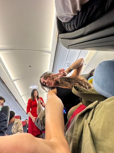 The image shows the interior of an airplane cabin, where a woman is taking a selfie. She is wearing headphones and making a peace sign with her fingers. There is a flight attendant in a red uniform visible in the aisle. The cabin is filled with passengers seated, some of whom are wearing masks. Overhead bins and the plane's ceiling are also visible.