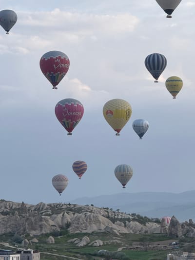 The image shows a clear sky filled with multiple hot air balloons, a popular tourist activity in the city of Göreme, Turkey. The landscape, visible on the right side, consists of unusual rock formations characteristic of the Cappadocia region. The buildings of Göreme are partially visible at the bottom right. No people are visible in the photo.