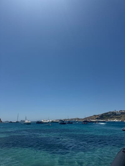 Azure sky dominates, meeting a strip of turquoise water crowded with anchored yachts and boats. A steep coastline dotted with buildings rises beside the water. The sea displays subtle ripples, reflecting light. A hint of a vessel is visible on the upper right corner.