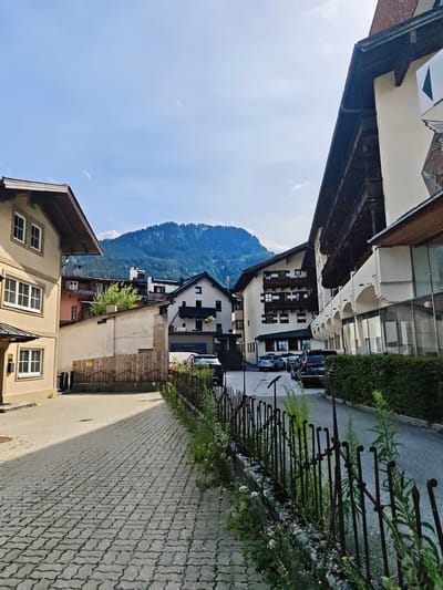 The image shows a street scene in Kitzbühel, Austria, with buildings on either side and a mountain in the background. On the left, a light-colored building with white-framed windows and a dark sloped roof is visible. A paved walkway made of interlocking grey bricks runs in the foreground, leading towards the buildings. A dark metal fence with ornate finials separates the walkway from a street on the right. Behind the fence, several cars are parked along the asphalt road, including silver and dark-colored SUVs. The buildings are multi-story structures with traditional alpine architecture, featuring dark wood balconies and white facades. In the distance, a large, densely forested mountain rises under a clear blue sky with a few wispy clouds. The overall impression is of a well-maintained, picturesque European town.