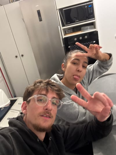 A selfie of two people indoors, rotated sideways. A young man with glasses and a goatee is in the foreground, wearing a black hoodie. Behind him is a woman with dark hair, sticking her tongue out and wearing a gray sweatshirt. Both are making hand gestures. A stainless steel refrigerator and white cabinets are visible in the background, along with a microwave and built-in appliances. The lighting is bright and cool.
