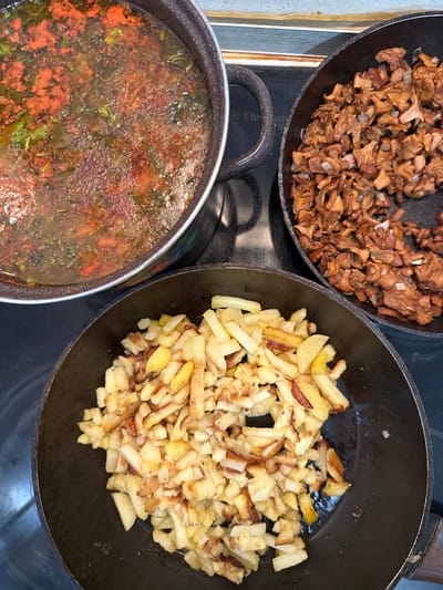 A close-up, overhead shot shows three pans on a black stovetop. In a large pot on the left, a reddish-orange soup with green herbs is simmering. To the right of the soup pot, a frying pan is filled with sautéed mushrooms and diced onions. Below these, another frying pan contains golden-brown fried potatoes, cut into strips and cubes. The overall scene suggests a home-cooked meal being prepared. The stovetop has markings suggesting it is a brand like Electrolux. There is no clear indication of the location within Sverdlovskiy, Russia.