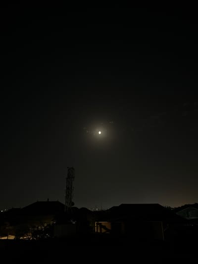 This is a nighttime photograph taken from an elevated vantage point, looking across a residential area in Sabunchu, Azerbaijan. The sky is dark, with a bright, luminous full moon positioned in the upper center of the frame. Wisps of clouds are visible around the moon, softening its glare.
In the middle ground, a tall, multi-tiered telecommunications tower stands prominently on the left side. Its metallic structure is silhouetted against the night sky. Below and around the tower, the dark shapes of rooftops from residential buildings are discernible. Trees with foliage are also visible in silhouette.
The foreground consists of dark, shadowy silhouettes of more rooftops, suggesting the observer is on a higher building or structure. In the distance, faint city lights are visible on the horizon, indicating the presence of a more urbanized area beyond the immediate residential neighborhood. The overall impression is one of a quiet, nocturnal scene in a suburban or semi-urban environment.