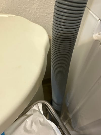 A close-up, eye-level, indoor shot depicts the narrow space between a cream-colored appliance and a wall. A flexible, ribbed gray hose runs vertically along the wall, occupying the center of the frame. To the left, a portion of the cream-colored appliance, possibly a washing machine or dryer, is visible with a smooth, curved surface. Below the hose and to the right of the appliance, a white plastic bag or laundry is partially visible within a chrome-rimmed basket. The wall behind the hose appears to have a textured, beige surface. The lighting is dim, casting subtle shadows. There is no indication of the city of Vancouver, United States of America, nor are there any people or actions depicted.