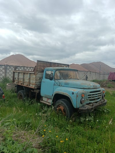 An old, blue, rusted truck sits in a field of green grass and dandelions on an overcast day in Яльгелево, Russia. The truck has a rusty metal bed and appears to be non-functional, with overgrown vegetation surrounding it. Behind the truck, a concrete fence borders a large pile of sand or gravel. Another vehicle, possibly a car, is partially visible on the left. The sky is filled with grey clouds.