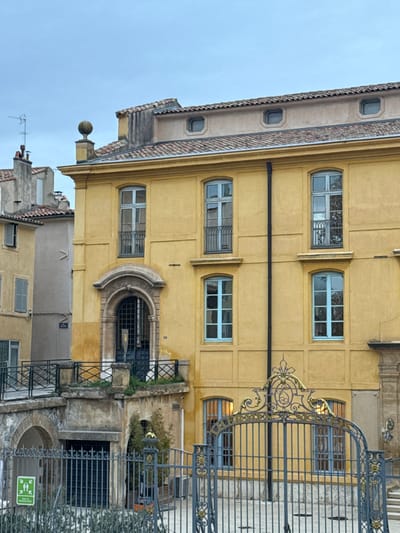 A yellow building with blue-framed windows stands tall. A wrought-iron gate with decorative flourishes obscures parts of the facade. The building features a stone archway and small balconies. A neighboring building is visible below, with tiled roofs and shutters. The sky is overcast.