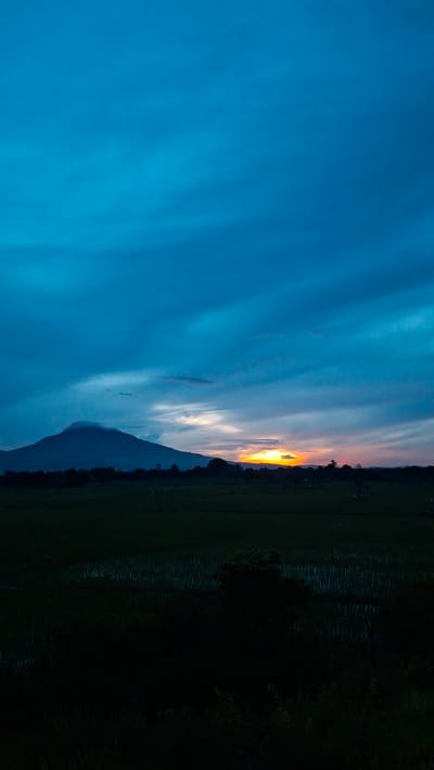 The image depicts a tranquil sunset scene over a rural landscape in Baksukon, Indonesia. The sky is a deep, vibrant blue with streaks of orange and yellow where the sun is setting behind a distant mountain range. The mountain, silhouetted against the colorful sky, is partially obscured by clouds. In the foreground, a dark, vast expanse of rice paddies stretches across the land, reflecting the dimming light. The overall impression is one of peacefulness and natural beauty, characteristic of the Indonesian countryside at dusk. There are no people or man-made structures clearly visible, emphasizing the natural and serene atmosphere of the location.
