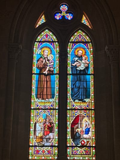 Two arched stained glass windows depict religious figures. The upper window shows a dark-robed figure holding a child, and another scene of kneeling and standing people with a book. The lower window portrays a brown-robed figure carrying a child, and a scene with people surrounding a figure in white. A smaller trefoil stained glass window is on the side, featuring a blue and red floral pattern. The surrounding architecture is grey stone.