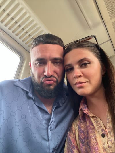 Two individuals, a man and a woman, are captured in a close-up, selfie-style photograph within what appears to be a train car. The man, on the left, has a beard and is wearing a blue collared shirt with a repeating pattern of interlocking "GG" logos. He is making a puckered facial expression. The woman, on the right, has long brown hair and is wearing a patterned collared shirt. Sunglasses are perched on her head. The background shows the interior of a train, including ventilation grilles and a window. The overall lighting suggests an indoor setting, possibly during daytime given the light from the window. The location is identified as the city of Izberbash, Russia.