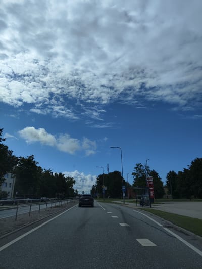 The image captures a street scene in Salacgrīva, Latvia, on a partially cloudy day. A dark-colored SUV is driving away from the camera on the right lane of a paved road. The road has white lane markings, including a solid white line on the right edge and dashed lines separating lanes. On the left side of the road, there is a metal fence separating the road from a sidewalk and a building. Trees and some parked cars are visible behind the fence. On the right side of the road, there are more trees, a bus stop shelter, and a red sign with white text that appears to be for a service station, displaying hours from 6-22. Streetlights are present on both sides of the road. The sky is a vibrant blue with scattered white and gray clouds.